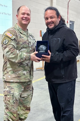 A man wearing a camouflage military uniform poses with another person holding a trophy.