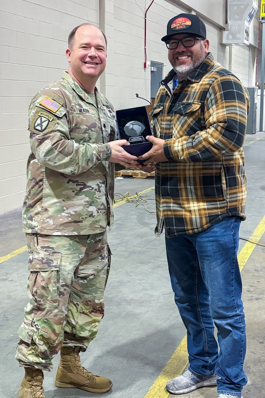 A man wearing a camouflage military uniform poses with another person holding a trophy.
