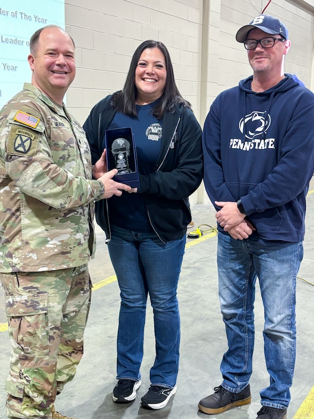 A man wearing a camouflage military uniform poses with two other people holding a trophy.
