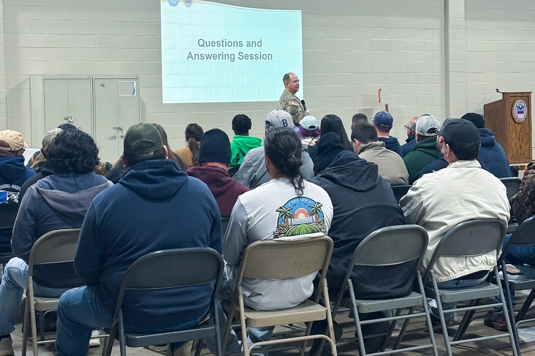 A man wearing a camouflage military uniform speaks to a crowd of people seated inside a large warehouse.