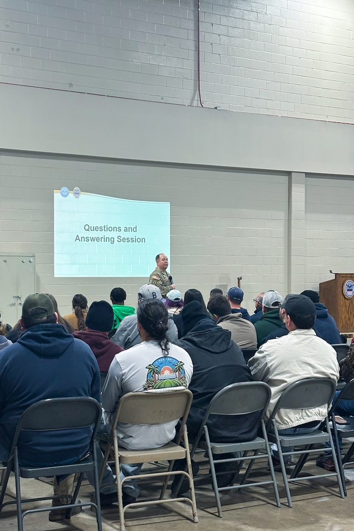 A man wearing a camouflage military uniform speaks to a crowd of people seated inside a large warehouse.