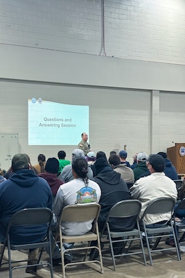 A man wearing a camouflage military uniform speaks to a crowd of people seated inside a large warehouse.