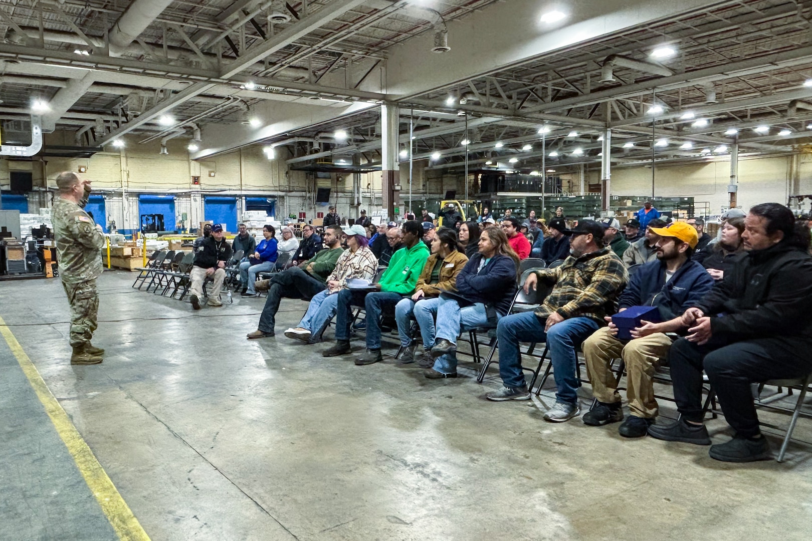 A man wearing a camouflage military uniform speaks to a crowd of people seated inside a large warehouse.