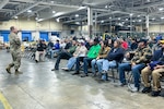 A man wearing a camouflage military uniform speaks to a crowd of people seated inside a large warehouse.