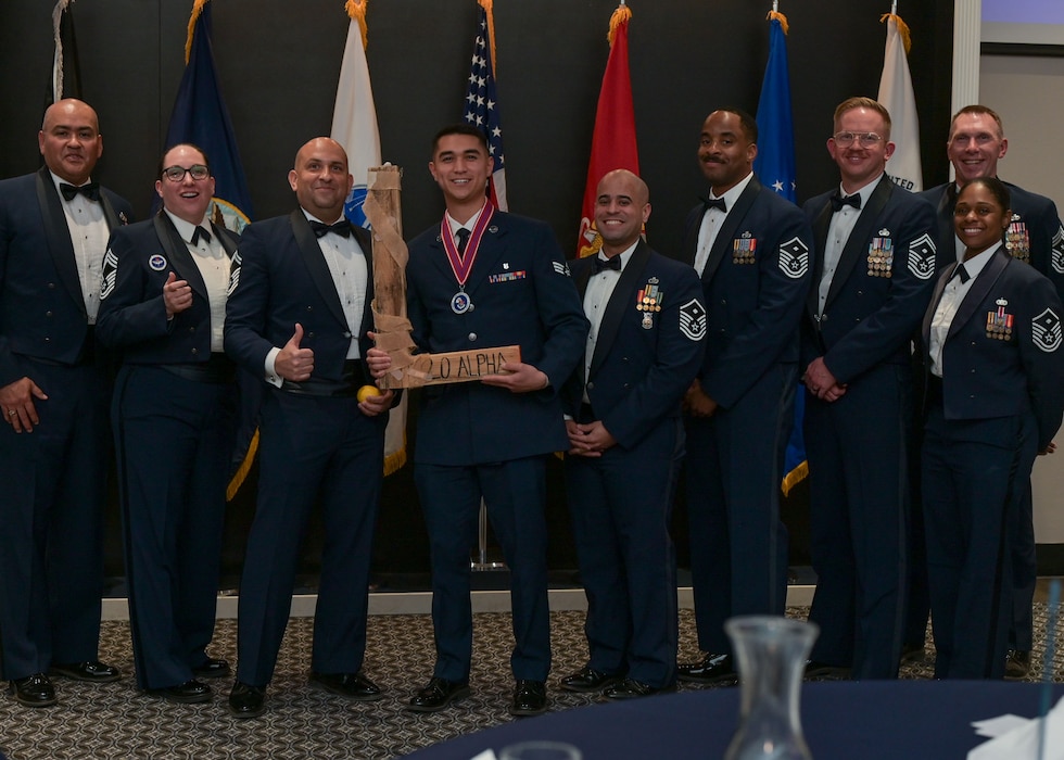 A wooden "L" is held up in the center of a group photo of Airmen from the ranks of senior airman to chief master sergeant.