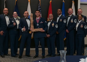 A wooden "L" is held up in the center of a group photo of Airmen from the ranks of senior airman to chief master sergeant.