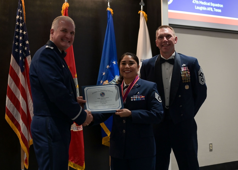 An Air Force colonel presents a graduation certificate to a senior airman as they pose for a picture together with a chief master sergeant.