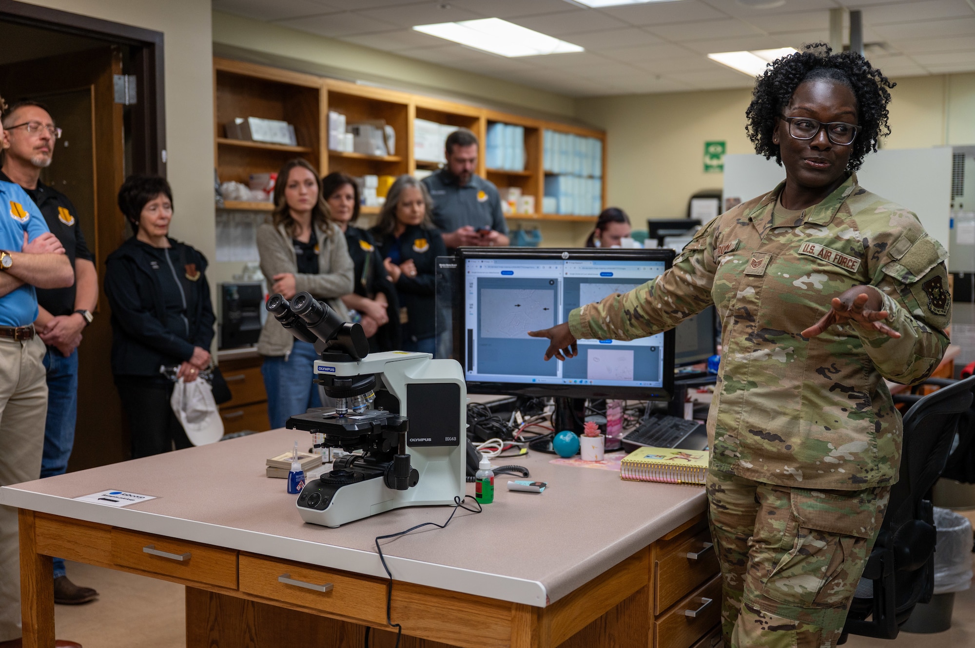A service member describes a microscopic sample shown on a monitor to a group of people.
