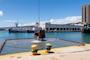 A clam shell bucket suspended by a chain is lifting out of water. In the background are two tugboats and a one-story harbor building.