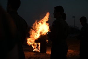 A piano engulfed in tall orange flames during a nighttime ceremony. The bright fire casts a warm glow, creating dark silhouettes of several people standing in a semi-circle around the instrument. The background is dark, emphasizing the sparks rising into the air and the contrast between the fire and the onlookers.