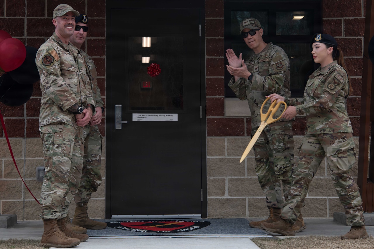 A woman in a camouflage military uniform is holding a giant pair of scissors after cutting a ribbon in front of a dog kennel facility. Three men in similar attire are standing in front of the building, with one of the men clapping his hands.