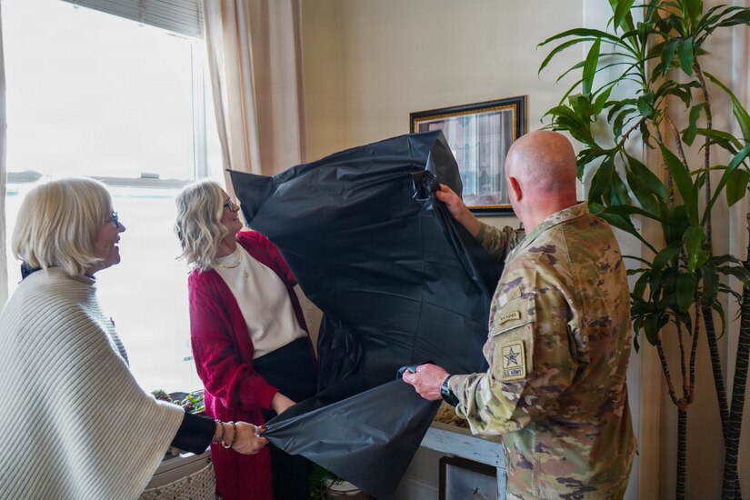 Two women in civilian clothes and a man in Army combat uniform are removing a black cloth from a framed picture on a wall during a dedication ceremony inside a house.