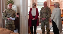 Army Soldier in combat uniform on left is gesturing while talking with two women in civilian attire and a man in combat uniform standing nearby inside a house.