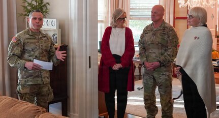 Army Soldier in combat uniform on left is gesturing while talking with two women in civilian attire and a man in combat uniform standing nearby inside a house.