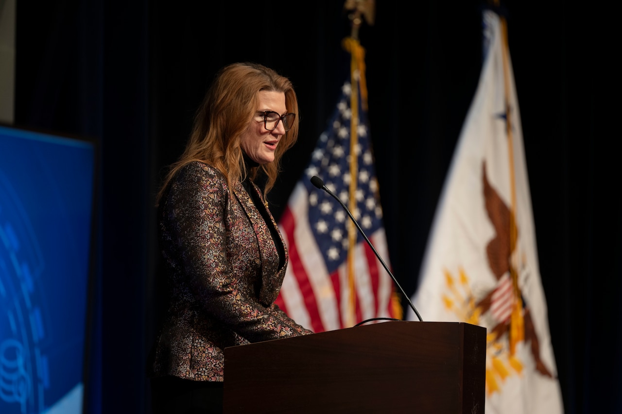 A woman in business attire stands behind a lectern and speaks into a microphone, with an American flag and a white flag with an eagle in the center behind her.
