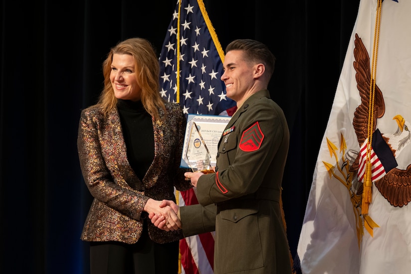 A woman in business attire presents an award to a man in a formal military uniform as they shake hands and smile to a camera off-screen; behind them are an American flag and a white flag with an eagle in the center.