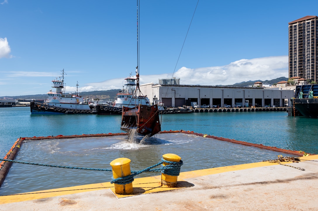 A clam shell bucket suspended by a chain is lifting out of water. In the background are two tugboats and a one-story harbor building.