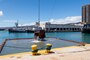 A clam shell bucket suspended by a chain is lifting out of water. In the background are two tugboats and a one-story harbor building.