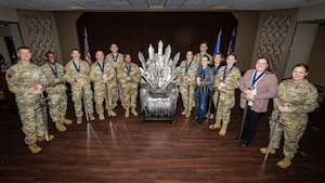 A group photo of military members, wearing camouflage uniforms, and civilians around a throne made of carboard swords
