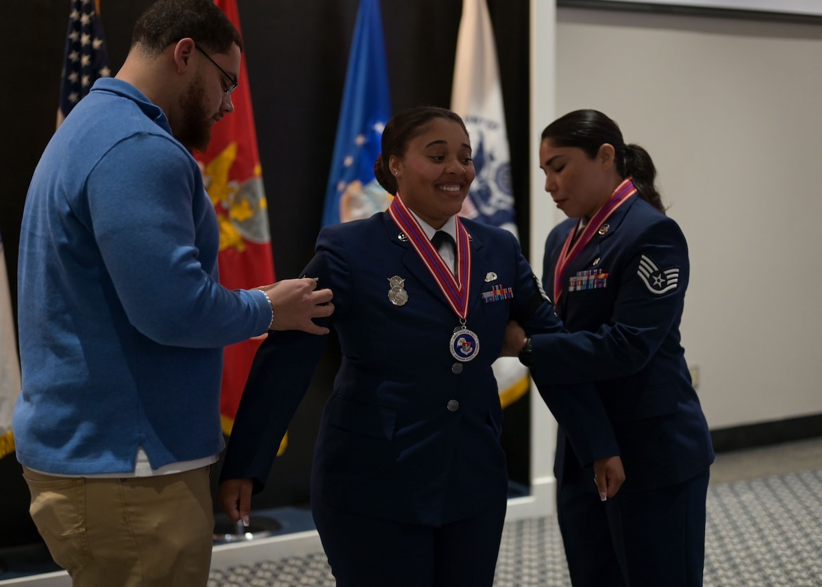 An Air Force senior airman stands with her arms out away from her body as her husband and colleague attach staff sergeant insignia to her arms.