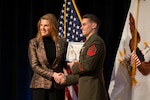 A woman in business attire presents an award to a man in a formal military uniform as they shake hands and smile to a camera off-screen; behind them are an American flag and a white flag with an eagle in the center.