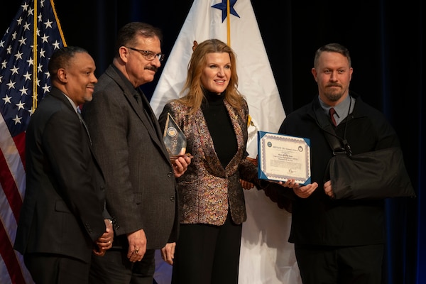 Three men and a woman, all in business attire, pose for a photo. One man holds a diamond-shaped trophy, and the woman holds a certificate; behind them are an American flag and a white flag with an eagle in the center.