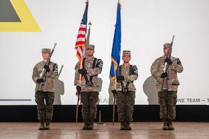 Four people stand on a stage, the two on the outsides hold parade rifles while the two in the middle hold the U.S. and Air Force flags