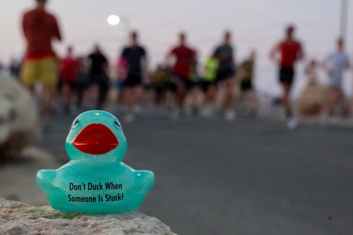 A rubber duck sits on a rock. On it is printed "Don't Duck When Someone is Stuck" People are blurred in the background
