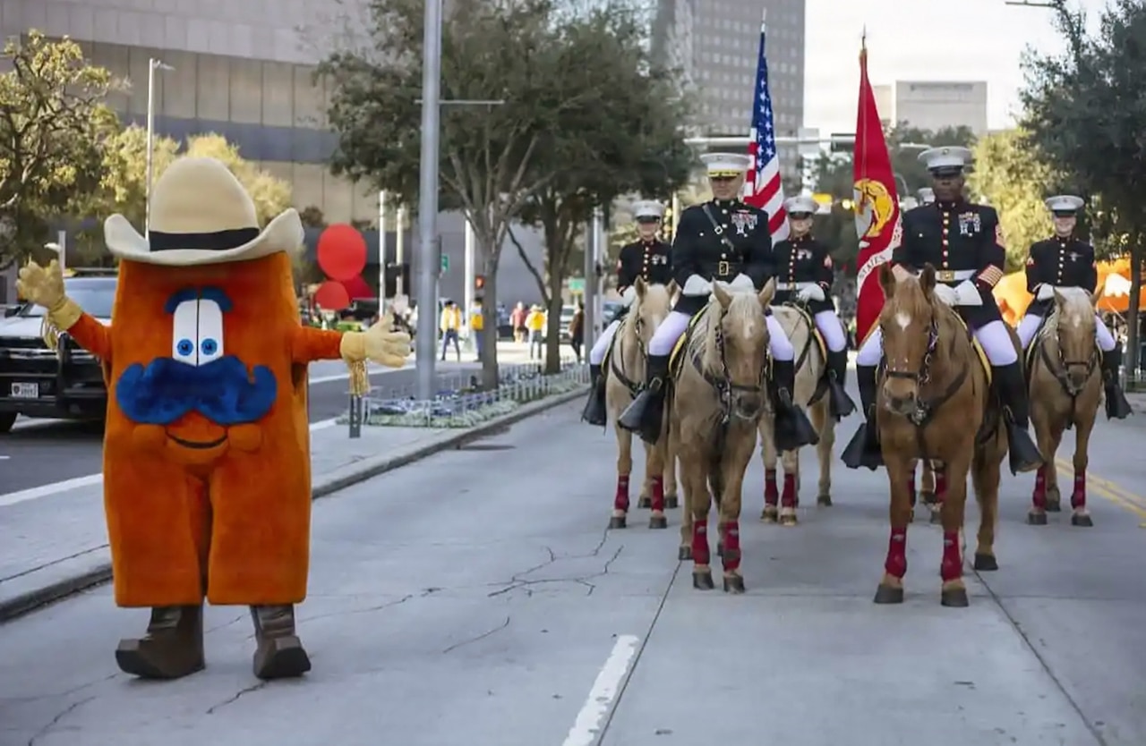 Six Marines in dress uniforms, two carrying flagpoles, ride horses in a parade procession near a cowboy-type mascot.