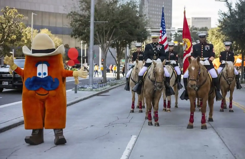 Six Marines in dress uniforms, two carrying flagpoles, ride horses in a parade procession near a cowboy-type mascot.