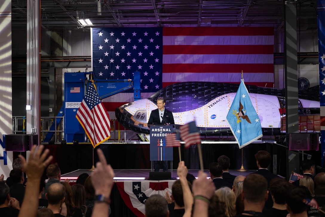 Secretary of War Pete Hegseth speaks at a lectern to a cheering audience with space equipment and a U.S. flag behind him.