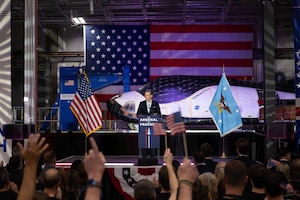 Secretary of War Pete Hegseth speaks at a lectern to a cheering audience with space equipment and a U.S. flag behind him.
