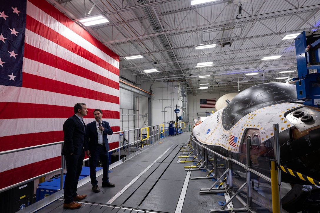 Secretary of War Pete Hegseth talks to a civilian while viewing a large piece of equipment in a large room with U.S. flag behind her.