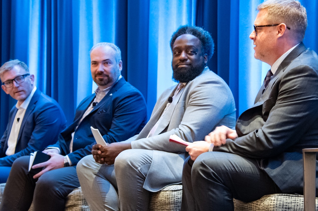 Four men are seated in chairs on a stage