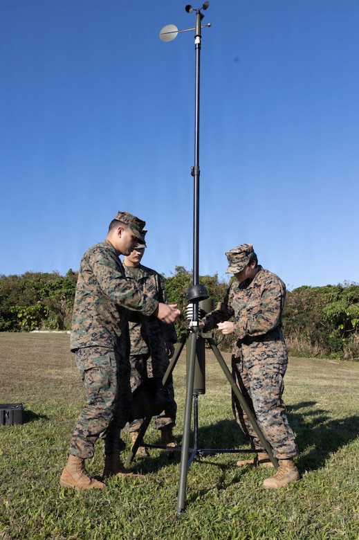 Three people in camouflaged military uniforms work on military equipment in a grassy field.