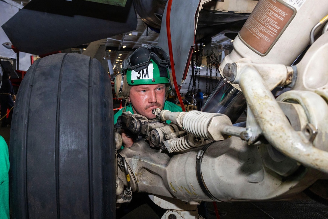 A man wearing a green helmet that says 3M on it looks through a maze of large pipes, with a large tire in the foreground.