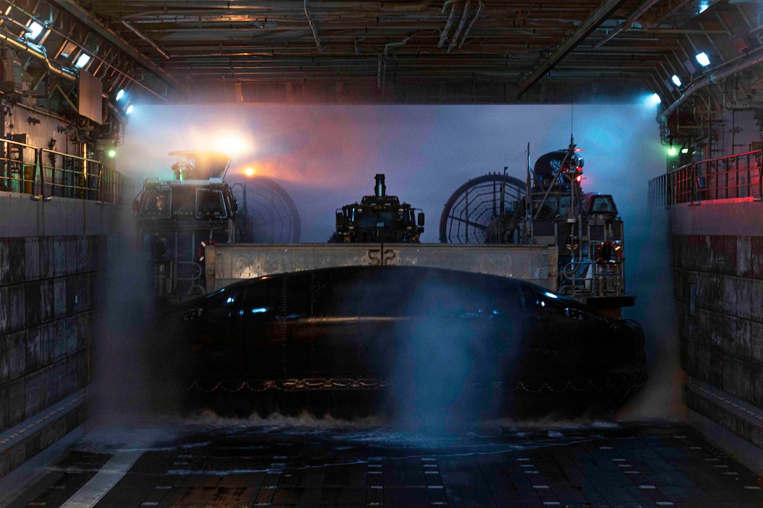 A military watercraft enters the well deck of a ship at night, with lights and mist behind it.