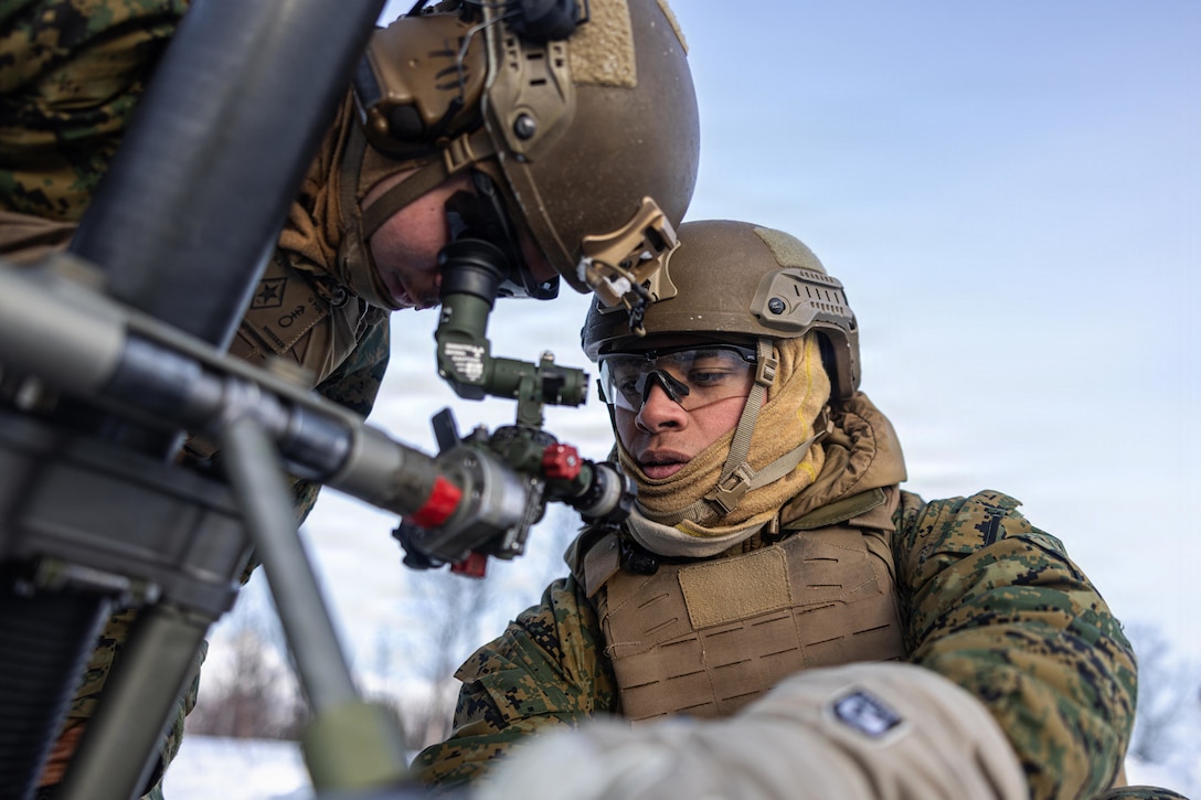Two people wearing camouflage military uniforms, helmets and safety goggles look down into a gauge, with a blue sky overhead and trees in the background.