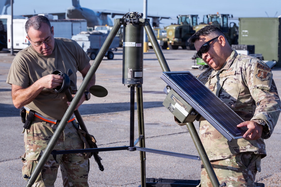 A man wearing a camouflage military uniform and sunglasses and a man in camouflage pants and a brown T-shirt assemble equipment in a parking lot, with vehicles in the background.