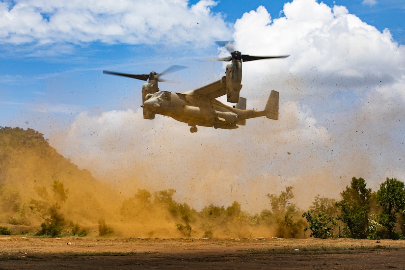 A tilt-rotor aircraft lifts off spreading dust through the air in a desert-like environment.