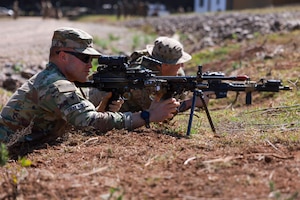 Two men wearing camouflage military uniforms point weapons at something outside of the photo frame while lying on the ground in the prone position.