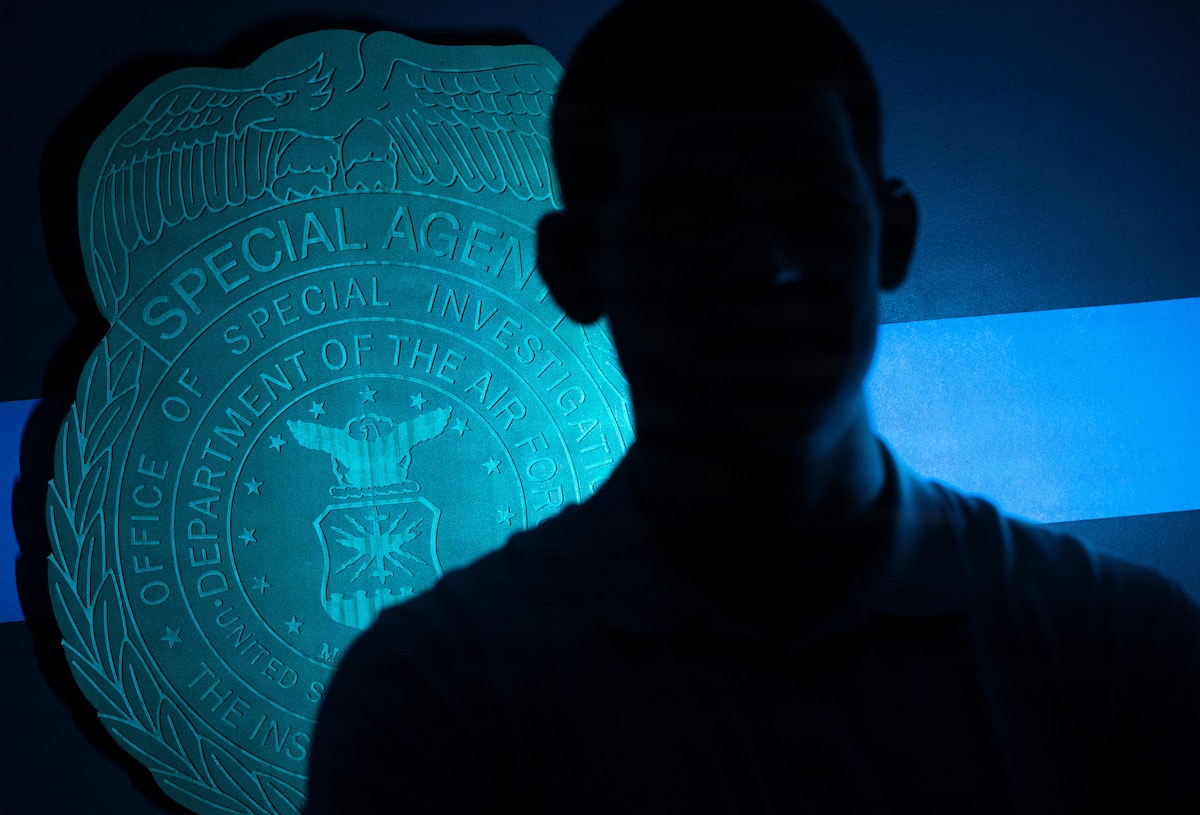 A U.S. Air Force Office of Special Investigations special agent stands near an AFOSI emblem and photos honoring fallen agents during a staged photo at Seymour Johnson Air Force Base, North Carolina, Feb. 11, 2026. AFOSI special agents serve worldwide to protect the Air Force and seek the truth in support of national defense. (U.S. Air Force photo by Airman 1st Class Holly Leonard)
