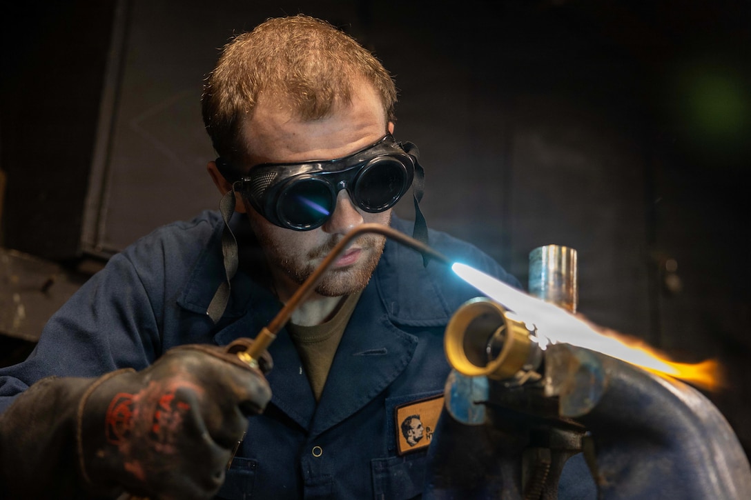 A man wearing a dark blue work uniform, welding gloves and safety goggles holds a torch and welds pipes, as white and yellow flames emit from the nozzle in a dark area.