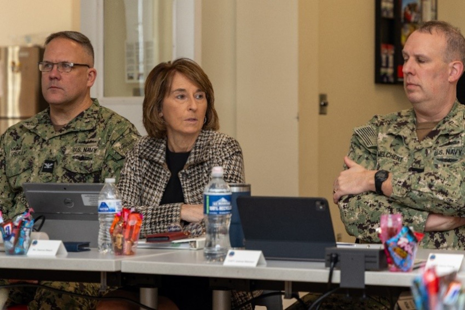 Woman with brown hair wearing brown, black and cream patterned jacket and black shirt under, sits with two men in military uniform.
