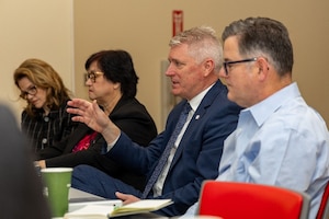Man with white/gray hair wearing blue suit gestures with right hand. Two women are seated to his right and one man seated to his left.