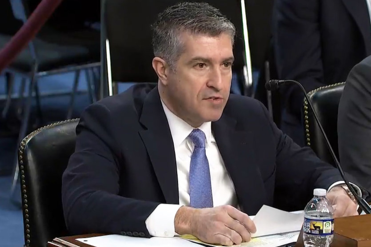 A man wearing business attire sits at a table and speaks into a microphone.