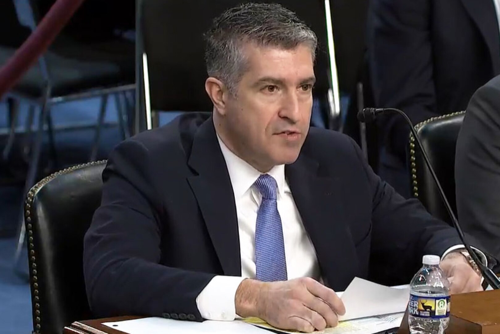 A man wearing business attire sits at a table and speaks into a microphone.