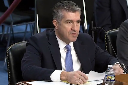 A man wearing business attire sits at a table and speaks into a microphone.