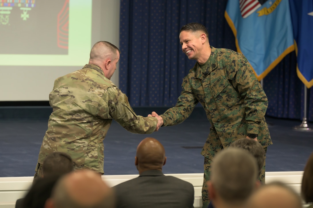 Two military servicemen wearing uniforms shake hands.