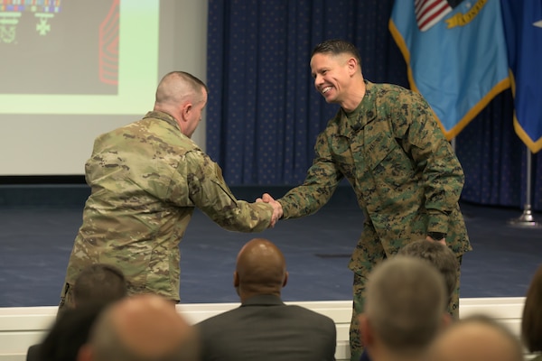Two military servicemen wearing uniforms shake hands.